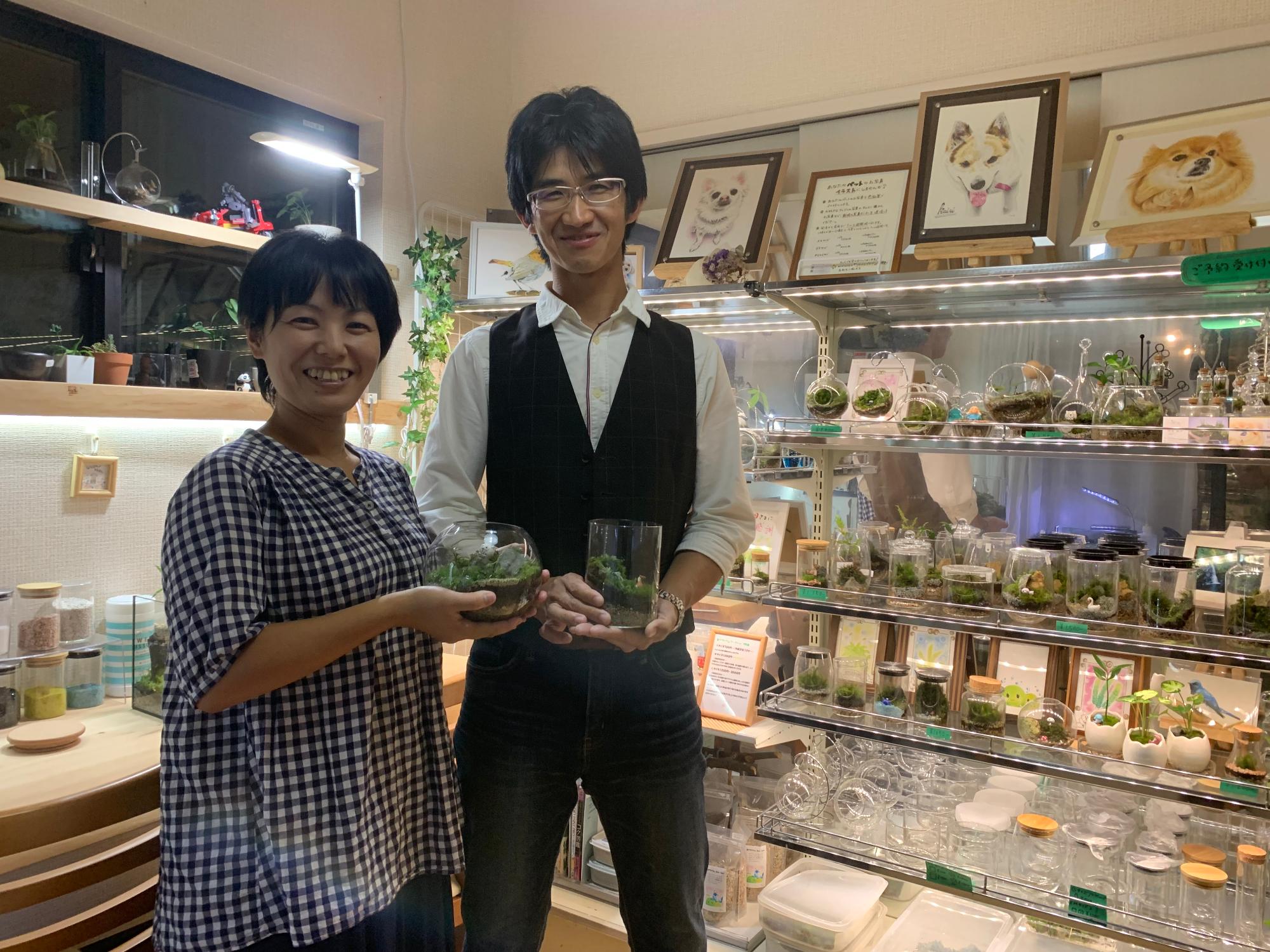 たくさんの苔テラリウムが並ぶ棚の前で、にこやかに立つ男女の写真