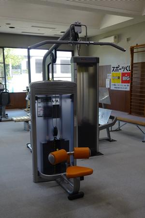 部屋の片隅に「スポーツくじtoto」の横断幕が貼られているトレーニング場で、様々なトレーニング機器に混ざってラットプルダウンというオレンジ色の椅子状のトレーニング機器が置かれている写真