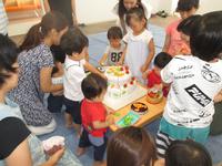 テーブルの上に置かれた白いケーキに子供達が囲むように集まり、すぐ後ろで保護者達が見守っている写真