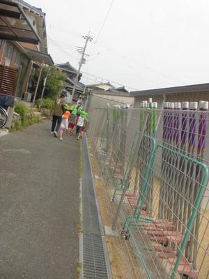 柵沿いの細い路地を歩いて行く子供たちと引率の先生の後ろ姿の写真