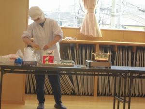 室内の長テーブルの上に並べられた食材を取り揃える、割烹着に衛生帽の先生の写真