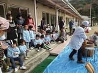 ブルーシートの上に置かれた臼と杵を使って餅つきをする女性2人を教室の前に座ったり立ったりして見守る児童たちと先生らの写真