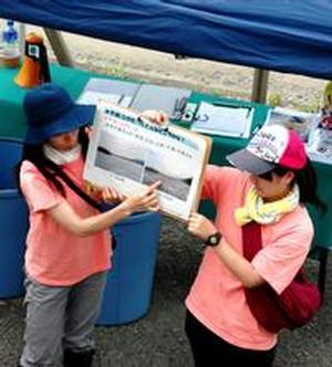 参加者たちが座る土手の階段下に設けられたイベント用テントの前でスタッフが持っているパネルを使いながら話をする前田真里さんの写真