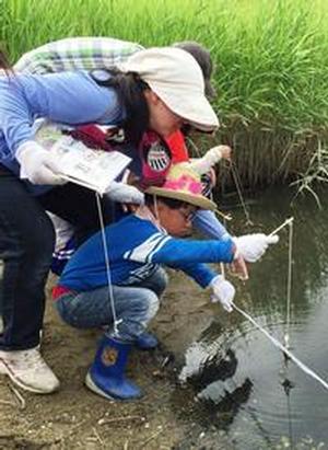 干潟の水辺にしゃがんで割りばしの先に紐を垂らしカニを釣ろうとしている子供たちと、その後で腰を曲げて前の子どもと同じ目線でそれを見守るスタッフの写真