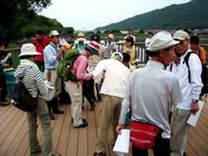 みどりの相談所前ウッドデッキスペースにて、小型の拡声器を手に参加者たちにイベントの説明をする松本先生と、それを聞く参加者たちの写真