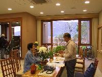 屋内でテーブルの上に花が複数置いてあり座っている人と立っている人がいて奥の大きな窓から大きな植物が見えている写真