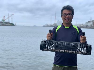 曇り空の海を背にして、両手で沢山の牡蠣が入っている網目状の筒を持って笑顔で写真に映るメガネを掛けた男性の写真