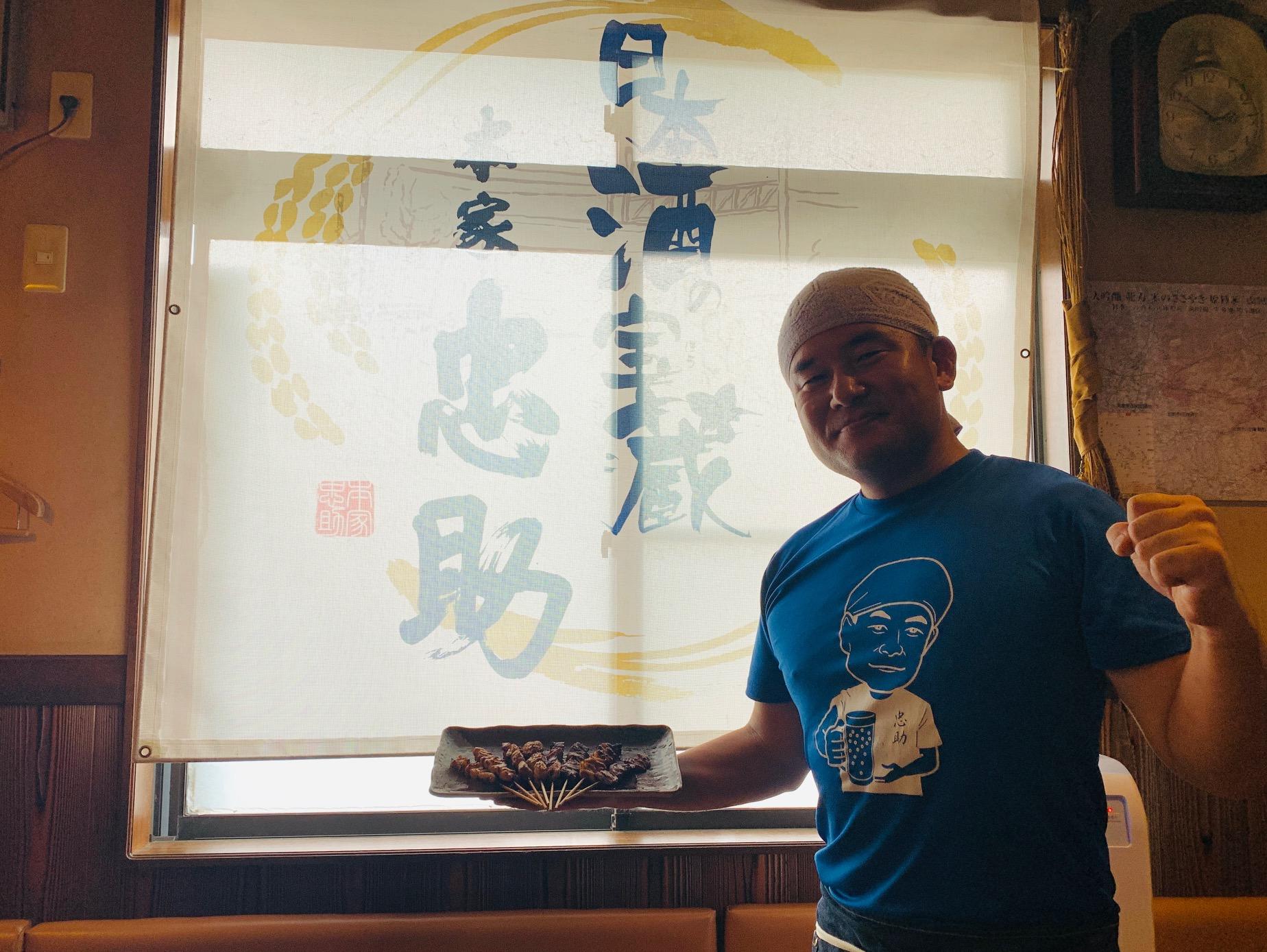 薄暗い部屋の中で、本家忠助と大きく書かれた垂れ幕の前で左手でガッツポーズをし、右手で串焼きの並んだ皿を持った笑顔の男性の写真
