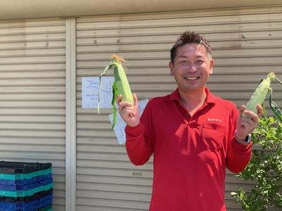 太陽が差し込む中、シャッターの降りた建物の前で赤いシャツを着た短髪の男性が笑顔でとうもろこしを両手で持っている写真