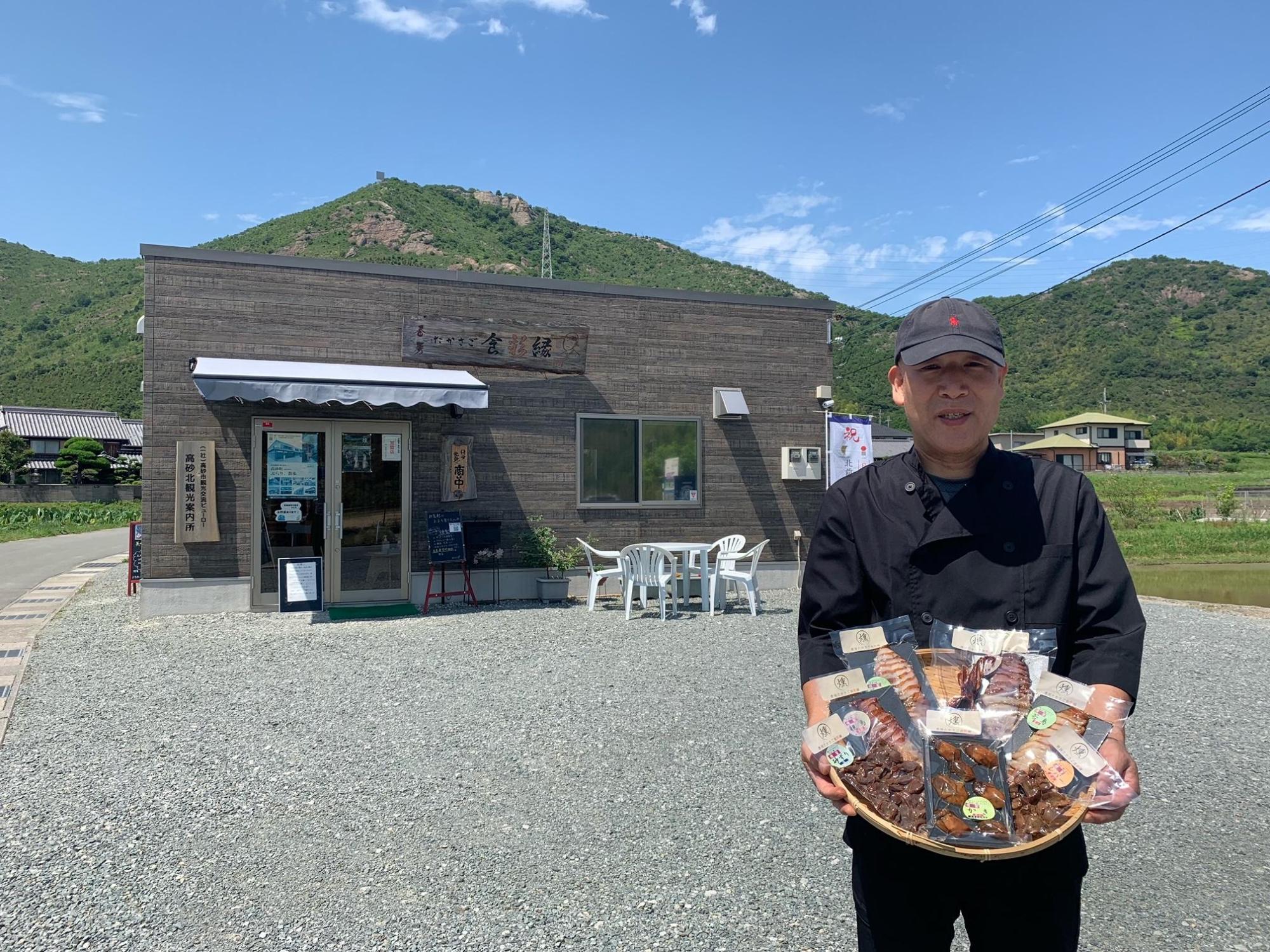 緑豊かな山に囲まれた風情ある木の壁で出来た店舗の前で、かごの入った商品を両手で持っている帽子をかぶった男性の写真