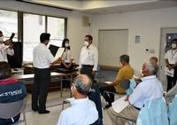 会議室のような室内で複数の男性が座って見守る中、1人の男性が表彰されている写真
