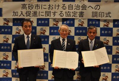 調印式でスーツ姿の男性3名が並んで証書を手にしている写真