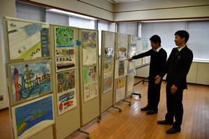 「高砂市の10年後」をテーマとして高砂市内の小学生が描いた絵画が高砂市役所南庁舎5階大会議室で展示され男性2人が間仕切りに貼られた数々の作品を眺めている写真