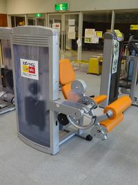 トレーニング場に置かれたレッグカール＆エクステンションというオレンジ色の斜め45度の座席で作られたトレーニング機器に「スポーツくじtoto」のロゴが貼られている写真
