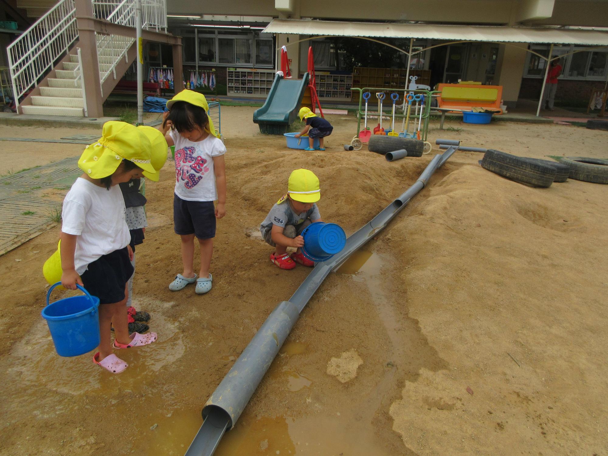 といを繋げて作ったトンネルにバケツで水を流す園児と足元に出来た水たまりを見ている園児3人の写真