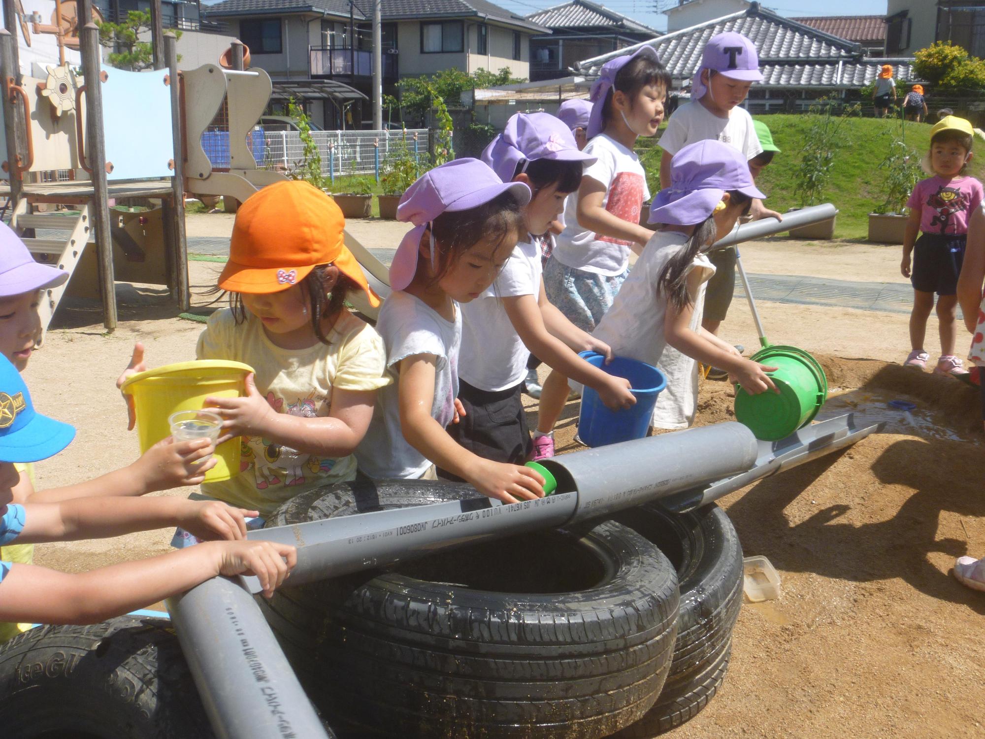 タイヤの上に置かれたトンネルにバケツで水を流す園児たちの後ろで水を流れやすくするためにといの位置を動かす園児たちの写真