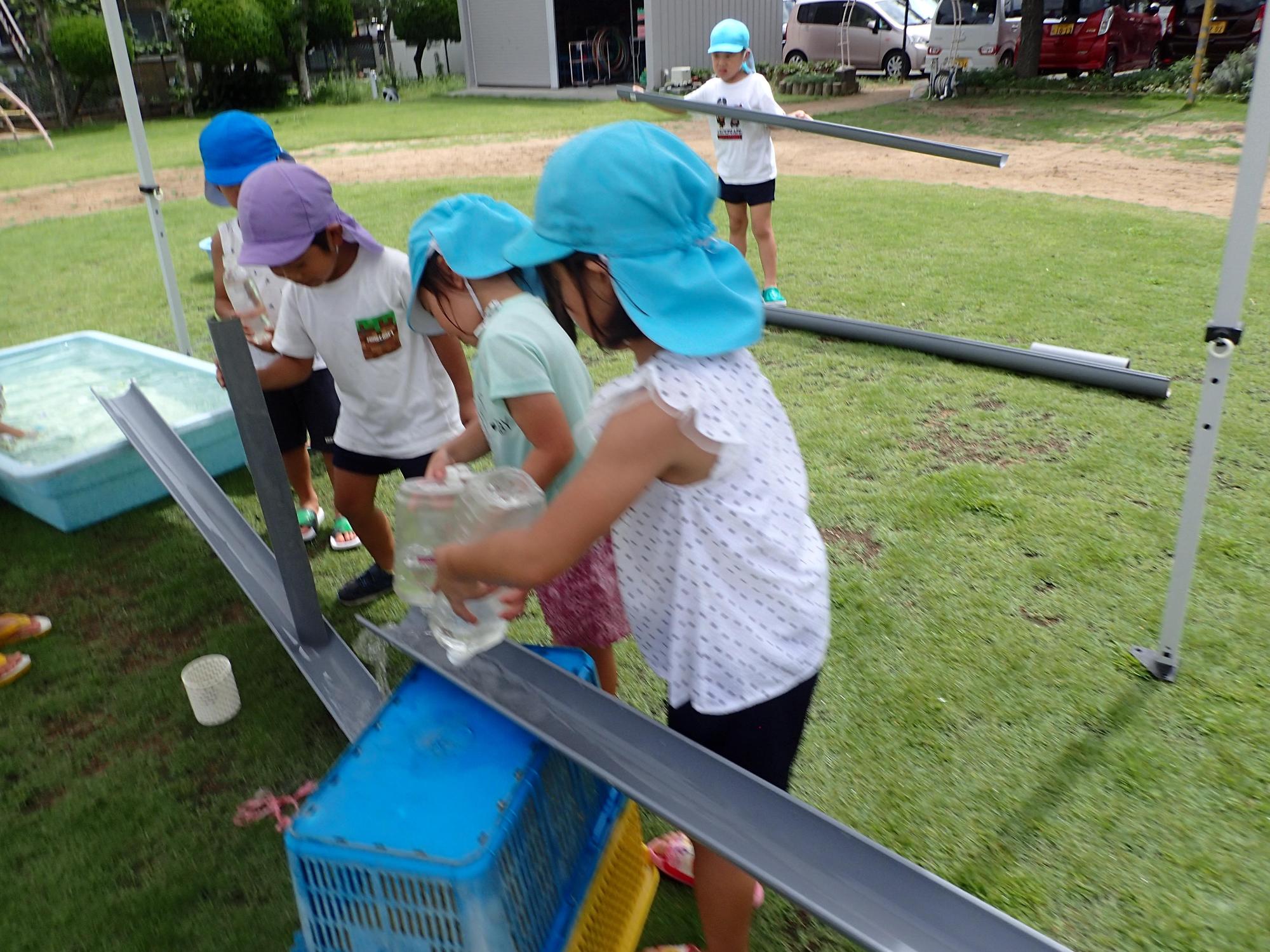 コンテナを土台にして置いたといにペットボトルに入った水を流す園児たちの写真