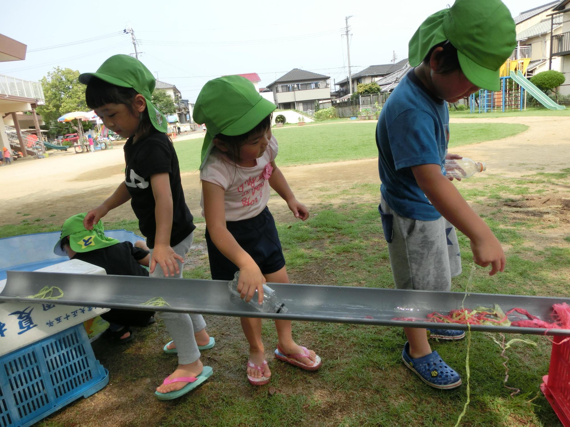 といにカラフルなそうめんを流していく男児の後ろでタイミングを合わせながらペットボトルに入った水を流す女児の写真
