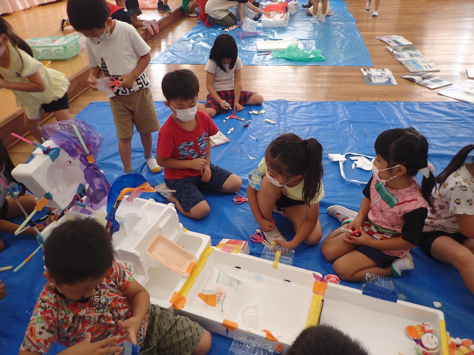 ブルーシートの上で廃材を使ってみんなで協力しながら船作りをする5歳児クラスの園児たちを正面から撮影した写真
