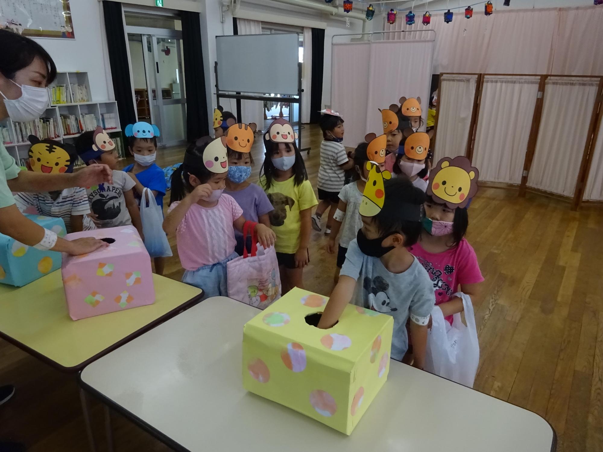 机に置かれた黄色のくじ箱の中に右手を入れている男児の様子を見守る園児たちと先生の写真