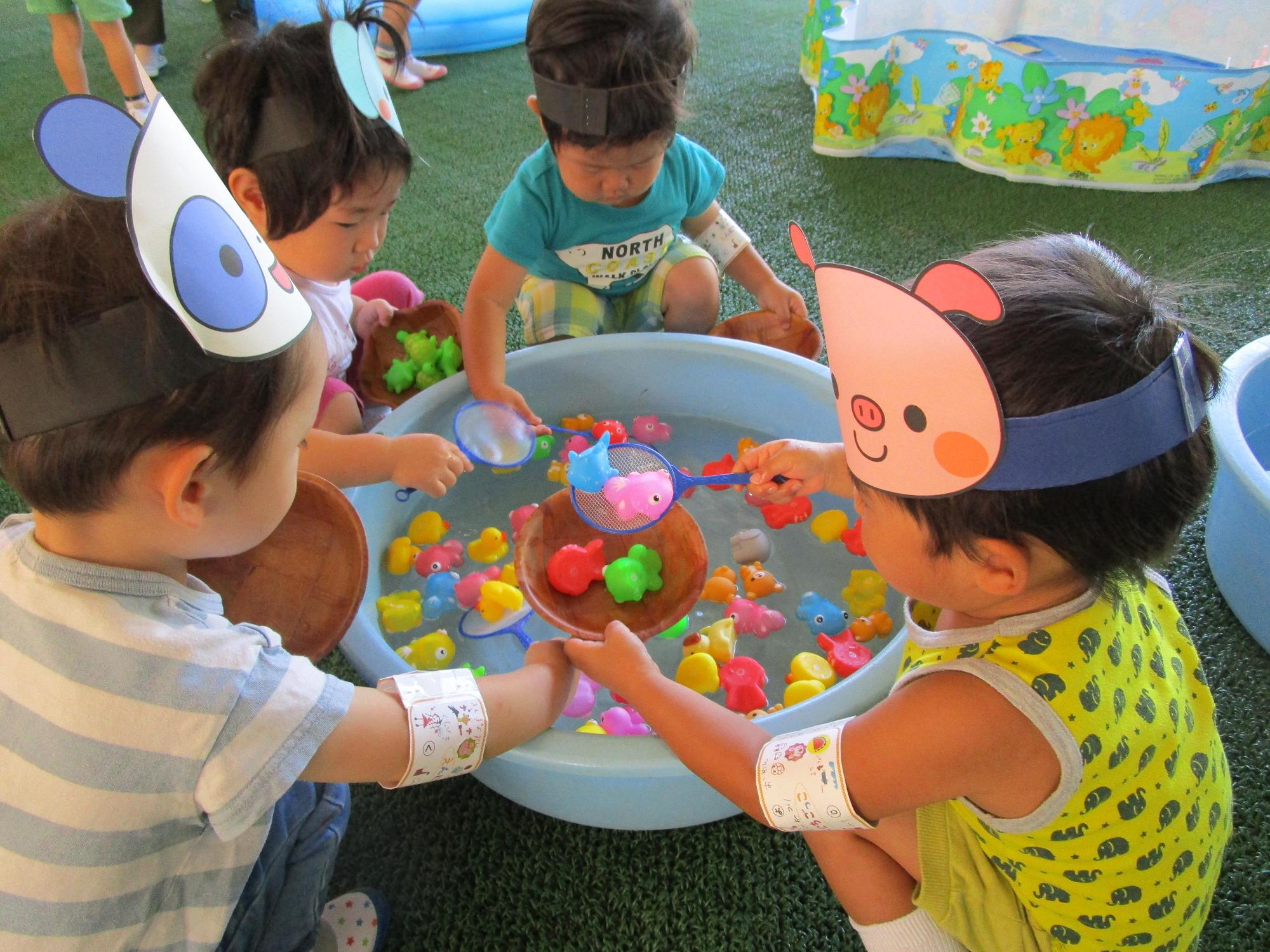 たらいに浮いているカラフルな金魚のおもちゃをポイですくってボウルに入れる頭に動物のお面を付けた園児4人の写真
