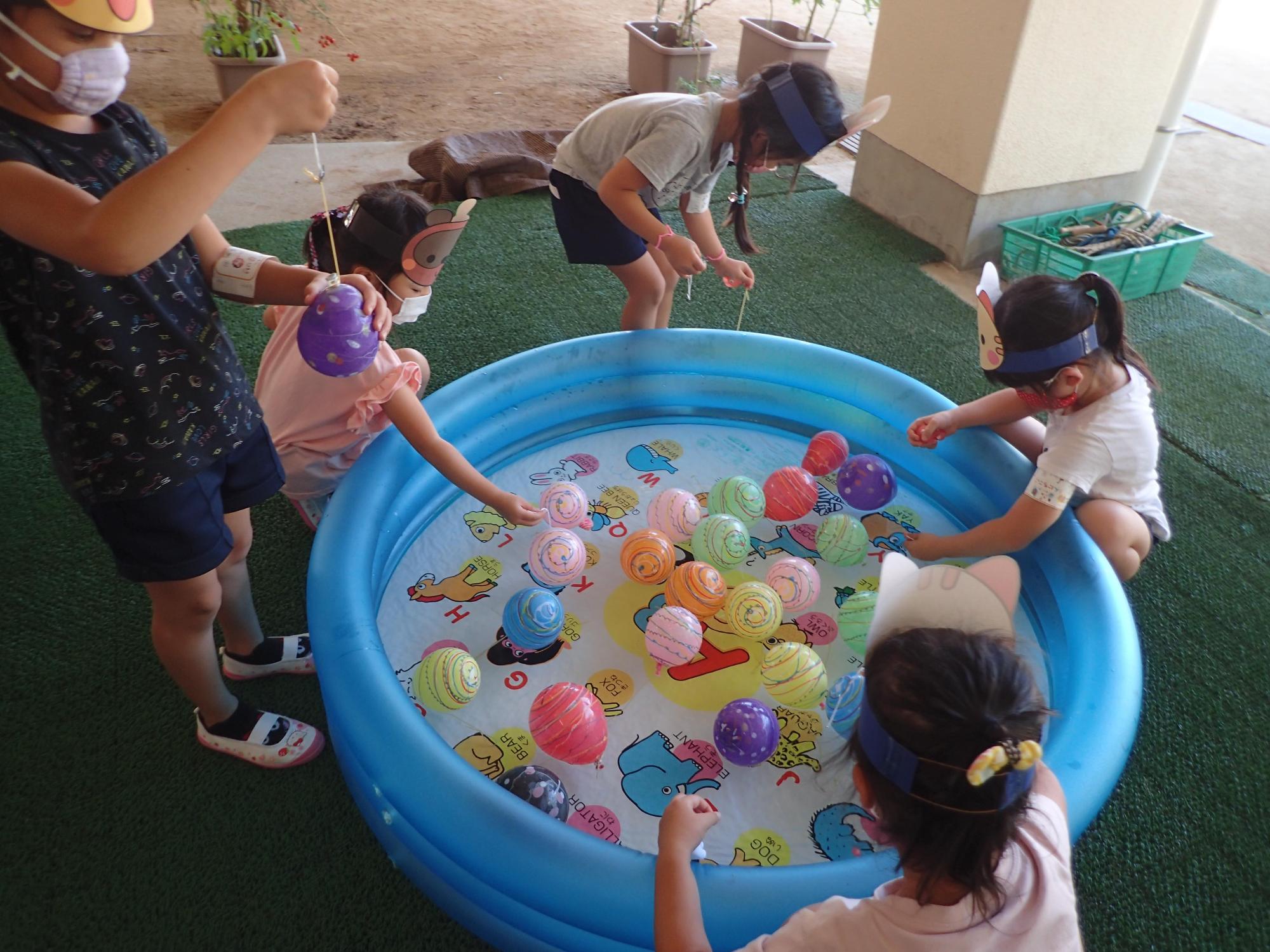 丸形のビニールプールに浮かんだヨーヨーを釣って遊ぶ頭に動物のお面を付けた園児たちの写真