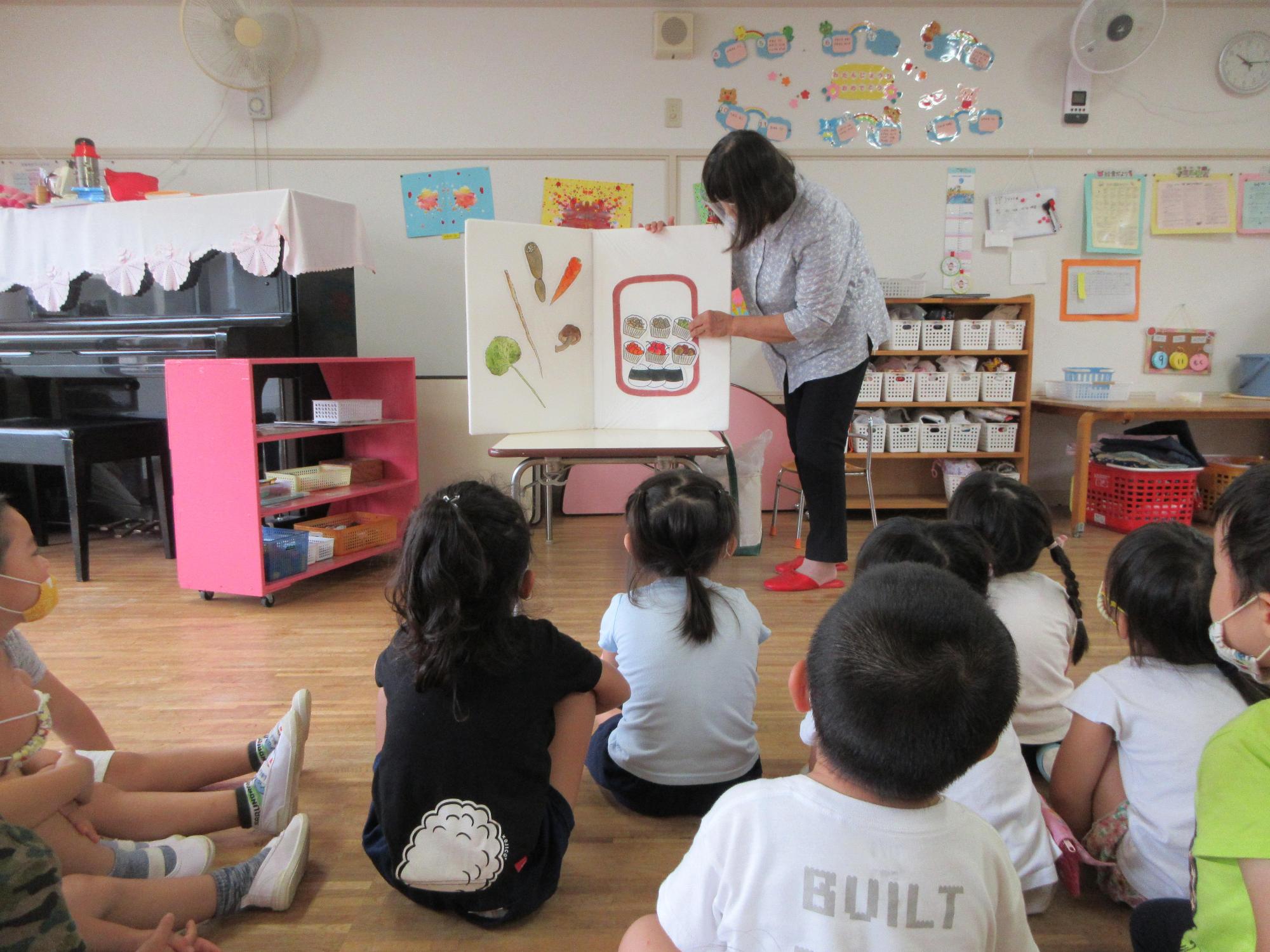 パネルシアターを披露する女性の方を体育座りをしたり足を伸ばしたりしながら見ている園児たちの写真