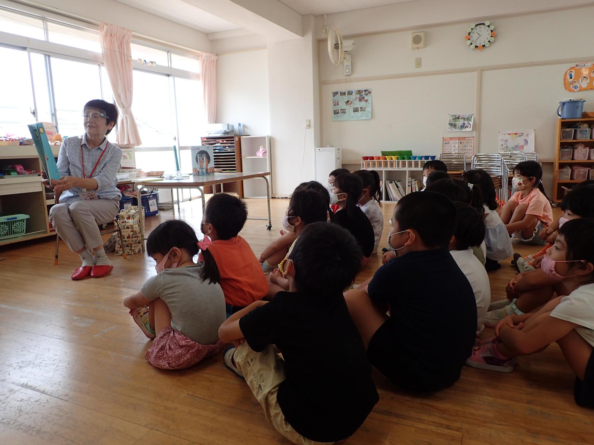 椅子に座って絵本を広げて読み聞かせをする女性の話を体育座りをしながら聞く園児たちの写真