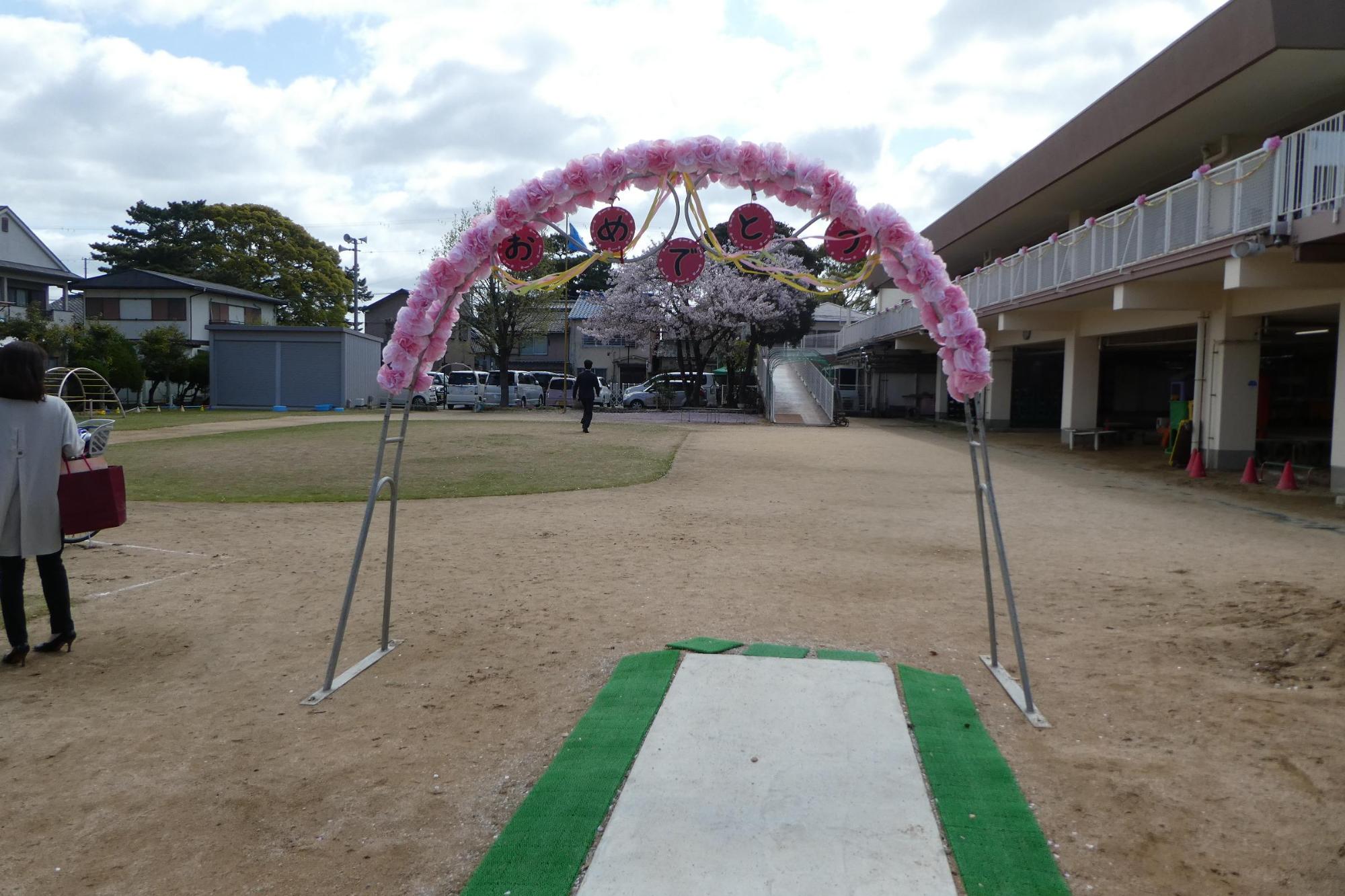 入園式に来る保育園児たちを迎え入れるために運動場にモール飾りやくす玉をつけたアーチがあり地面に緑と白のシートが敷かれている写真
