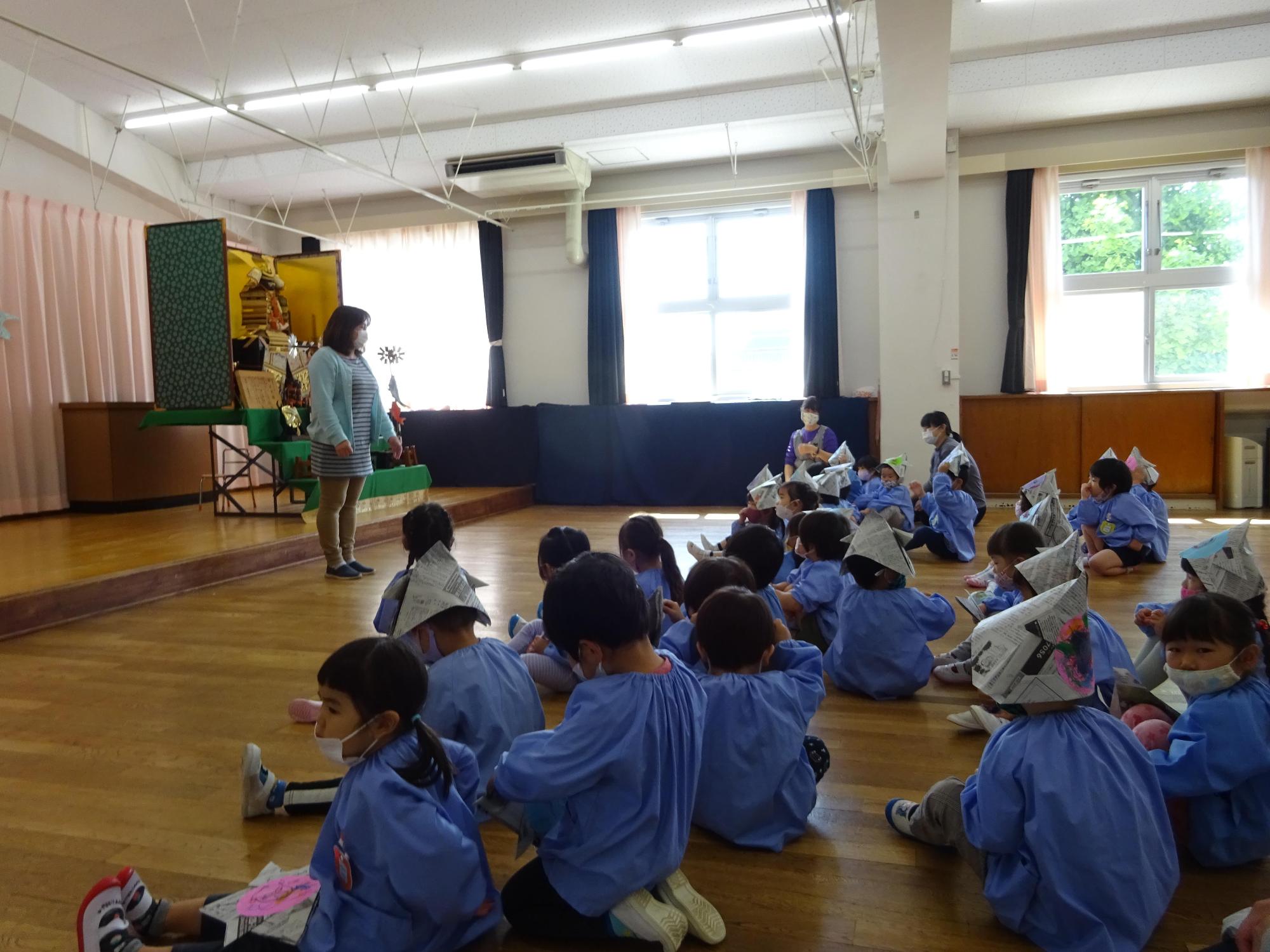 フローリングの大きな部屋に集まり座っている保育園児たちと「こどもの日」に関するお話をしている園長先生の様子を全体的に撮影した写真