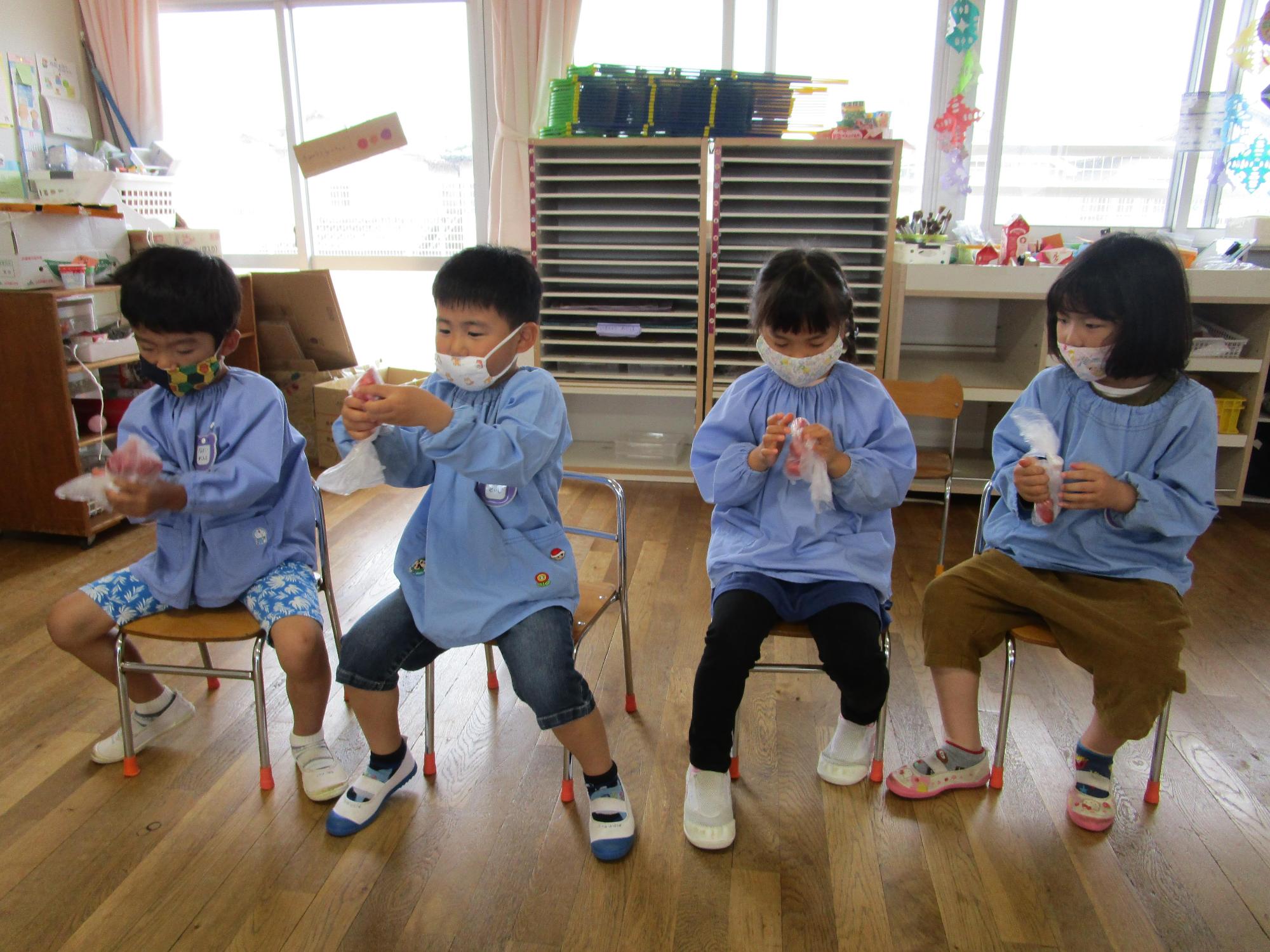 4名の保育園児が椅子に座りナイロン袋に入れたいちごを手で潰していちごジャムを作っている写真