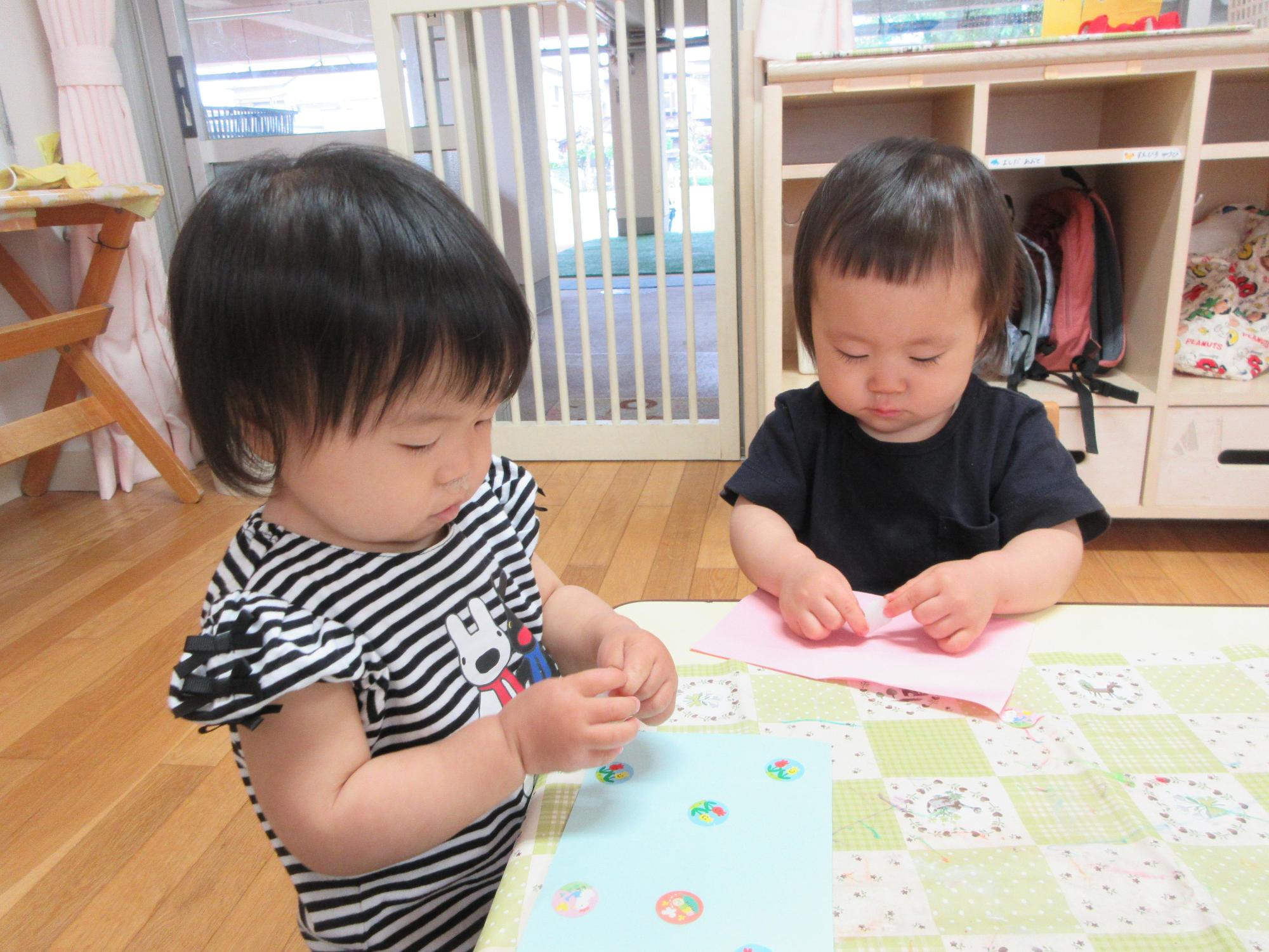 2人の保育園児が室内で赤い紙や青い紙にシールを貼っている様子の写真