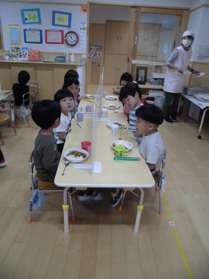 感染防止のパーテーションをはさんで机を挟んで食事をする子供たちの写真