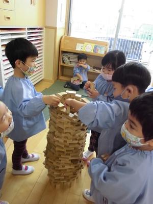 木のブロックを使って子供の背ほどある円筒状の塔をつくる子どもたちの写真