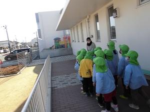 緑色の帽子をかぶりながら列になって並び、教師の交通安全の話を聞く園児たちの写真
