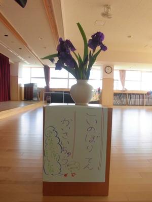 菖蒲の花が生けられている花瓶の下に、「こいのぼりてんかいさいちゅう」と書かれた手書きポスターが貼られている写真