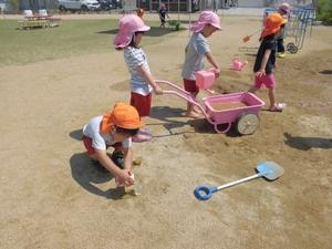 園庭の砂の上で、手押し車やじょうろなどを手に砂遊びをしている子供たちの写真