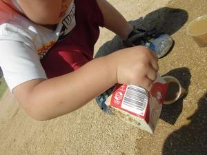 園庭の砂の上に置かれたカップに、牛乳パックを傾けて中に入った水を注ぎ入れる子供の写真