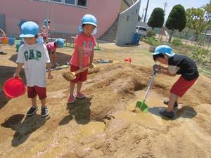 園庭の砂をシャベルで掘り、穴に水を流し入れて遊ぶ子供たちの写真
