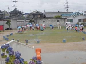 雨空の下、アジサイの咲く園庭を傘を差しながら歩く先生と子供たちの様子の写真