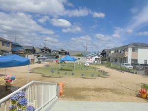 白い雲が浮かぶ青空の下、広々とした園庭に置かれている青い日よけテントとその周りに並べられたバケツの写真