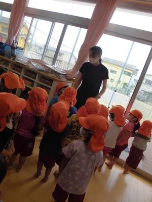 室内で立っている先生の周りに集まる、帽子を被って準備している子供たちの写真