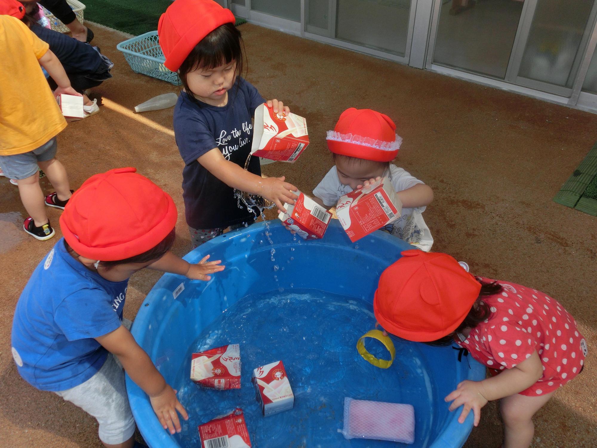 たらいに浮かんでいる上半分がカットされた牛乳パックを手に取り思い思いに遊ぶ園児たちの写真