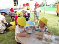 耳がついた黄色のニット帽を被った園児が氷の塊を眺めている左横から氷を触ろうと右手を伸ばす同じ帽子を被った園児の写真