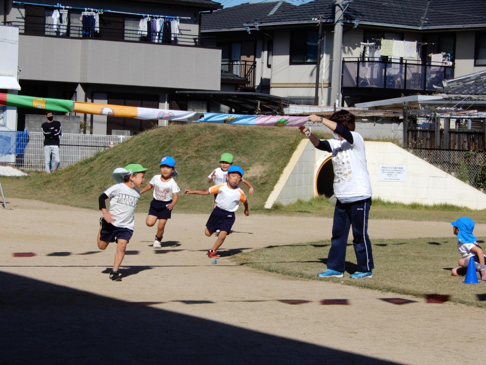 かけっこで先生の掲げているゴールテープに向かって走る園児たちの写真
