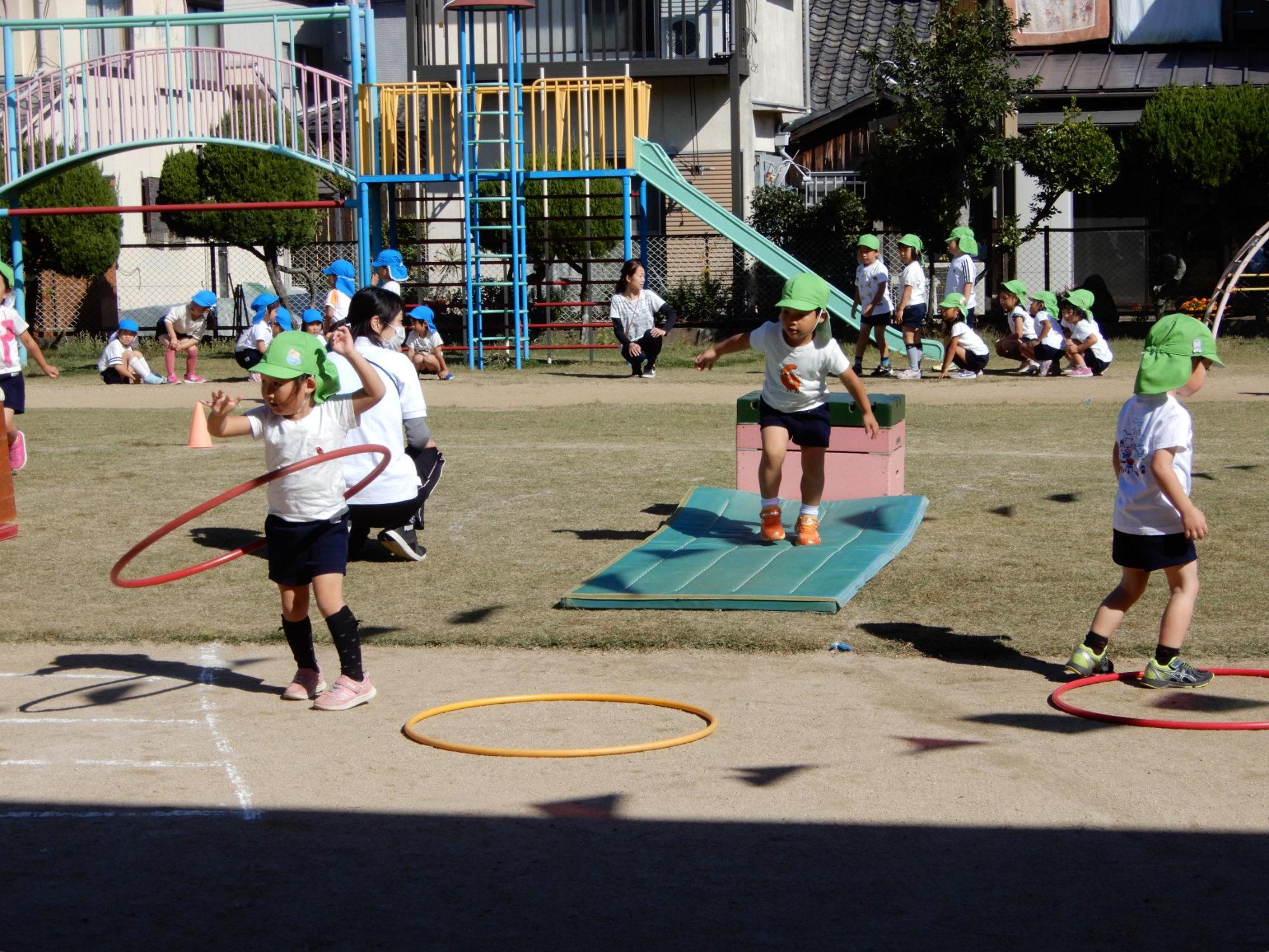 障害物競走でマットの上を走り次の障害物であるフラフープ方へ向かう園児の前でフラフープを回している園児の写真
