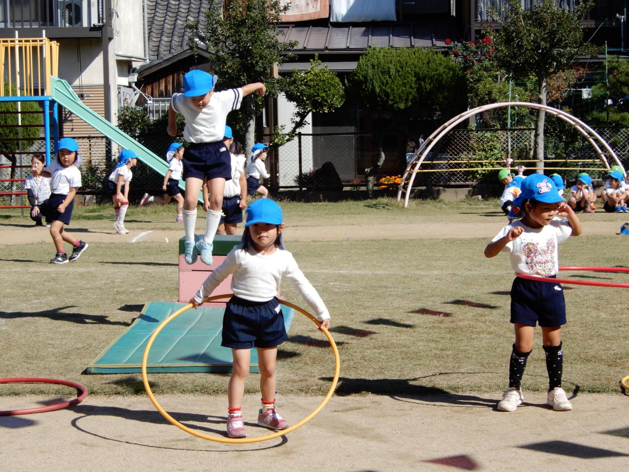 障害物競走で1段の跳び箱を飛んでマットに着地しようとする園児の前でフラフープを回している園児の写真