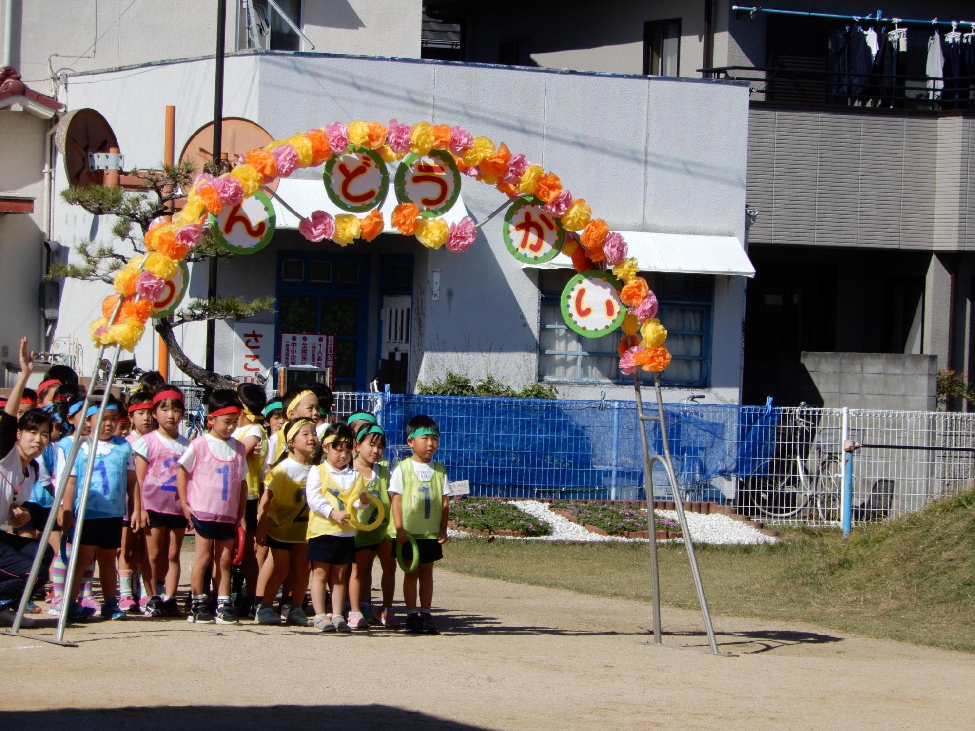 丸紙に一文字ずつうんどうかいと書かれた花アーチの下で整列する園児たちの写真