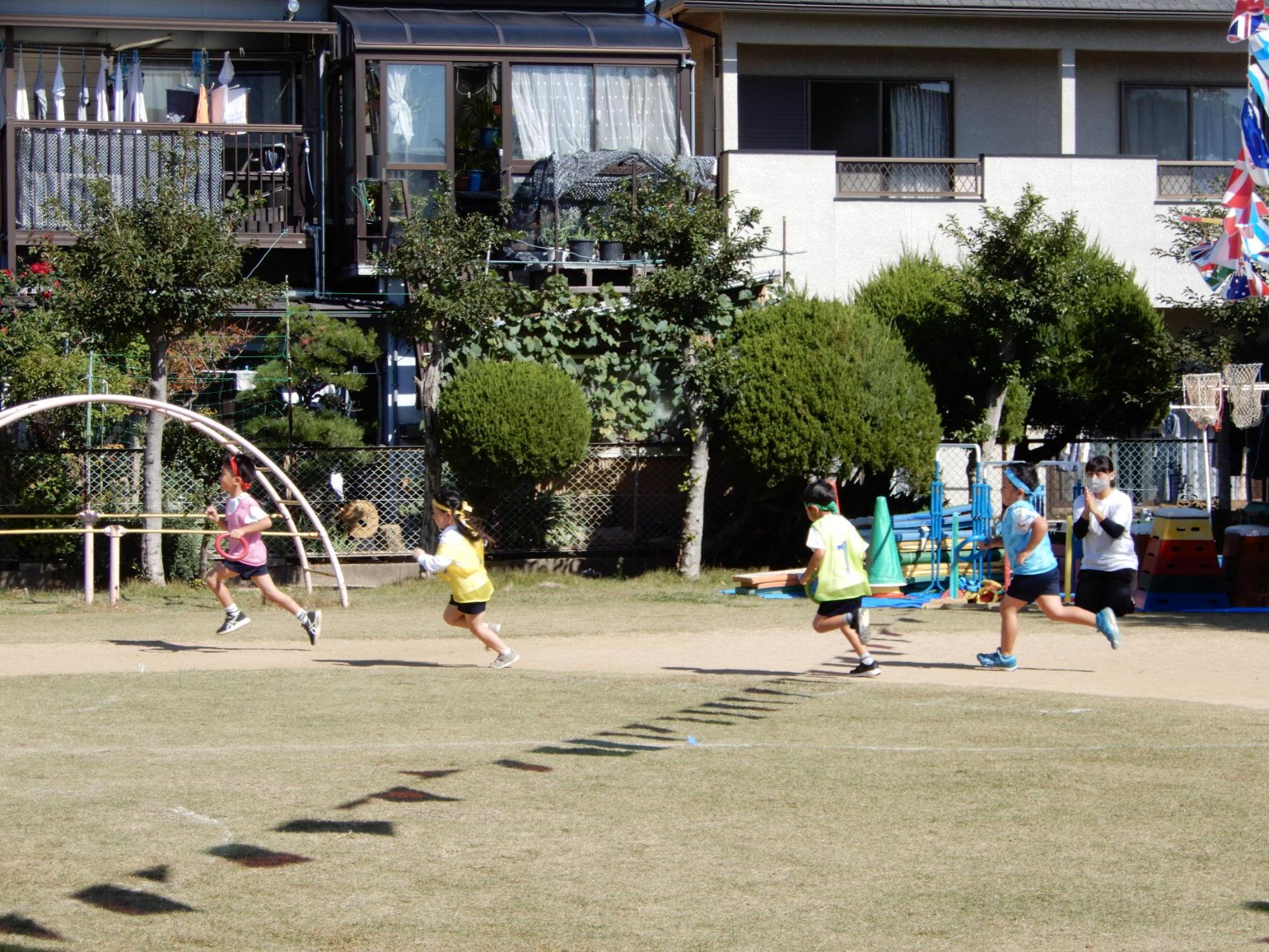 組の色のはちまきとぜっけんをしてリレー競争をする園児たちを手を叩いて応援する先生の写真