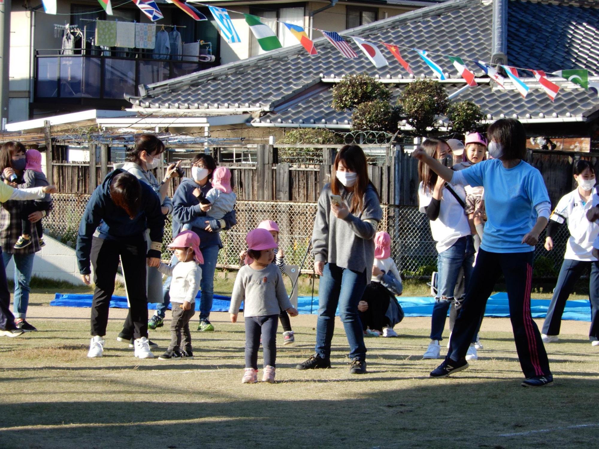 万国旗の下で先生の動きに合わせて保護者に抱っこされたり手を繋がれたりして踊る園児たちの写真